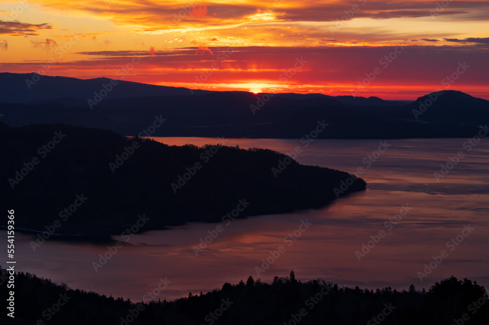 太陽光にオレンジ色の染まる空の色を弱く反射する湖。山と島の影。日本の北海道の美幌峠から見下ろす屈斜路湖。