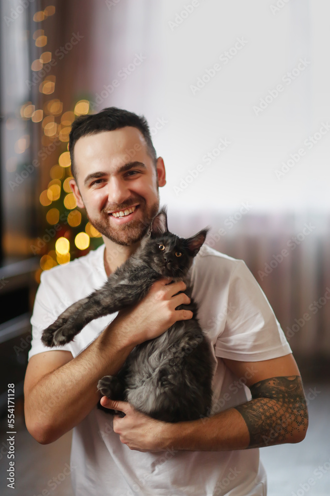 Obraz premium Portrait of manlike young man with beard with kitten in his arms. Kitten of Maine Coon kitten breed in arms of male owner. Christmas tree. Cozy Christmas time.