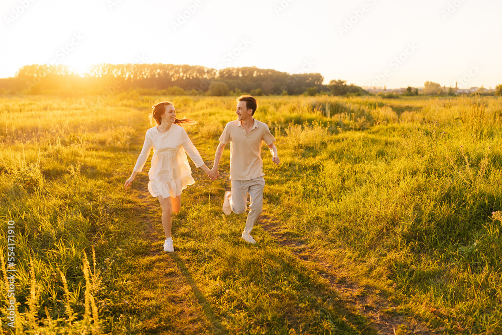 Happy young couple in love running looking at each other in green filed during golden sunset on summer sunny evening. Loving man and redhead woman embracing on background of bright sunlight. Happy young couple in love running looking at each other in green filed during golden sunset on summer sunny evening. Loving man and redhead woman embracing on background of bright sunlight.