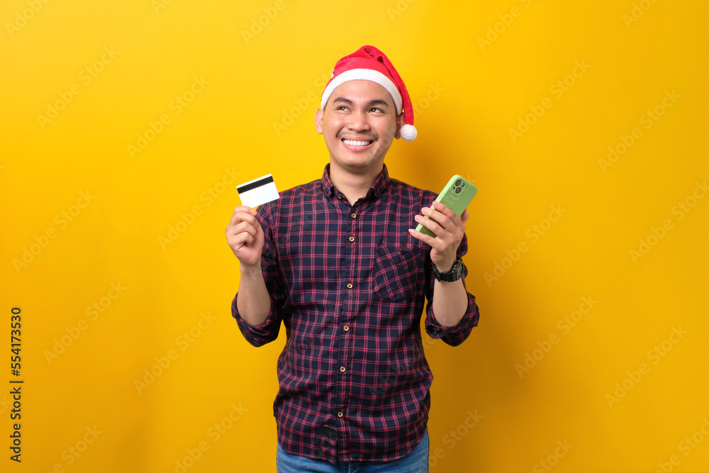 Smiling young Asian man in Santa hat using mobile phone and credit card, looking up, over yellow studio background. Happy New Year 2023 celebration merry holiday concept