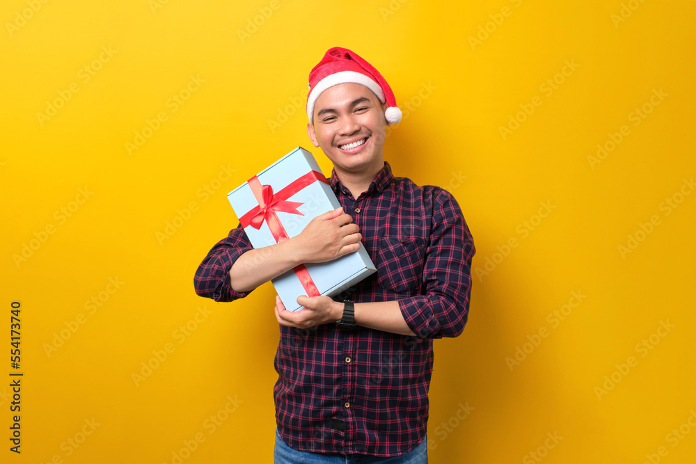 Smiling young Asian man in Santa hat holding gift box with ribbon on yellow studio background. Happy New Year 2023 celebration merry holiday concept