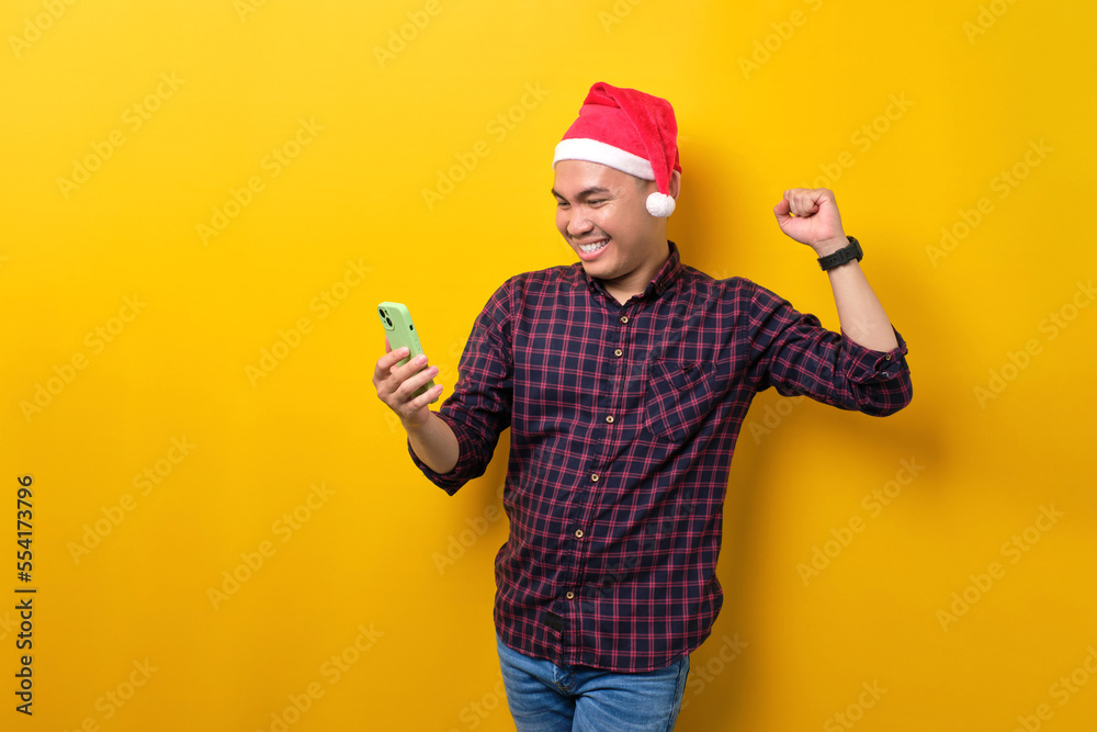 Excited young Asian man in Santa hat using smartphone and doing winner gesture over yellow studio background. Happy New Year 2023 celebration merry holiday concept