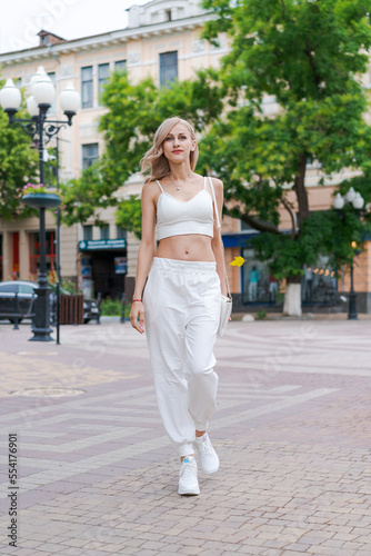 Happy young beautiful woman walking down street. Portrait cheerful university student looking at the camera, wearing white street clothes. Caucasian stylish girl smiling while standing.