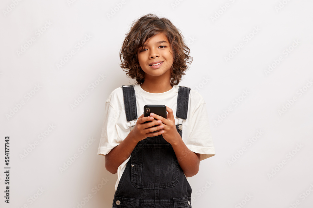 Charming young guy with curly hair, holds smartphone and send text messages, smiles gladfully, wears casual cloth, isolated over white wall Charming young guy with curly hair, holds smartphone and send text messages, smiles gladfully, wears casual cloth, isolated over white wall