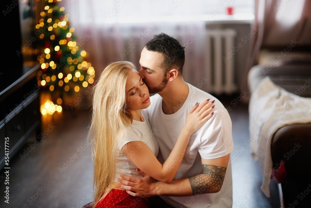 Young man tender kiss woman hugging on living room floor. couple in their red pajamas in living room by Christmas tree. Cozy good Christmas morning. Woman with big lips and long hairs. Young man tender kiss woman hugging on living room floor. couple in their red pajamas in living room by Christmas tree. Cozy good Christmas morning. Woman with big lips and long hairs.