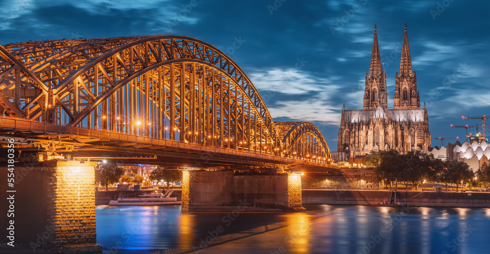 Panorama of illuminated bridge over Rhine river whith trains and tourists passing by and the Cologne Cathedral in the evening at the blue hour.