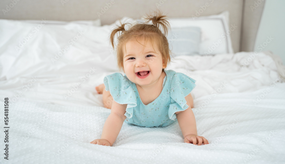 Happy baby girl smiling in blue suit pajama in white bedroom lying on bed looking at camera. Cute child with pigtails. Childhood, babyhood, people concept. Copy space fro advertisement Happy baby girl smiling in blue suit pajama in white bedroom lying on bed looking at camera. Cute child with pigtails. Childhood, babyhood, people concept. Copy space fro advertisement