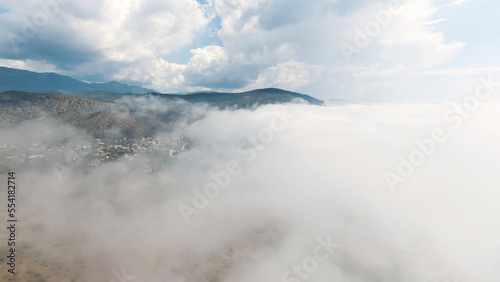 Top view of thick clouds with mountain peak on horizon. Schot. Flying in cloudy sky with dense clouds from top of mountain. Landscape of top of green mountain on horizon among white clouds
