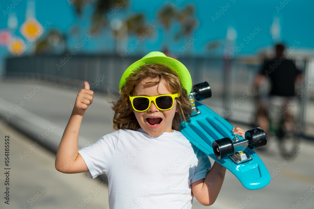 Fototapeta premium Skateboarder child boy in skate park. Kid boy with skateboard. Excited child with thumb up, street portrait close up. Caucasian fashion kid with bright facial emotions. Childhood, expression, having
