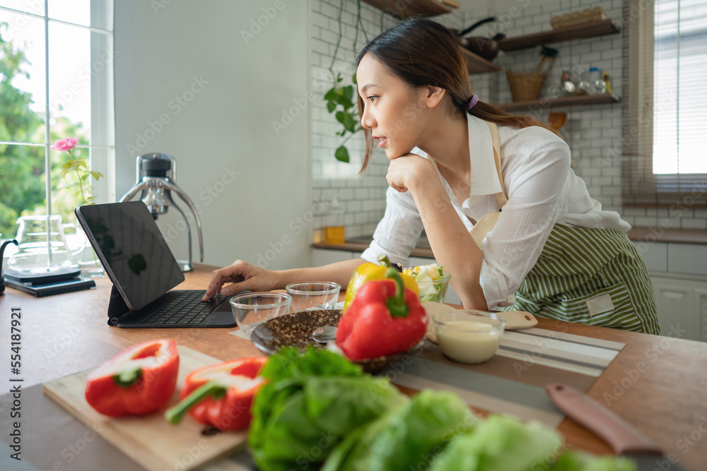 Asian housewife cutting fresh vegetables mixing salad ingredients cooking tasty vegan meal look search online blogger recipe notebook stand kitchen indoors