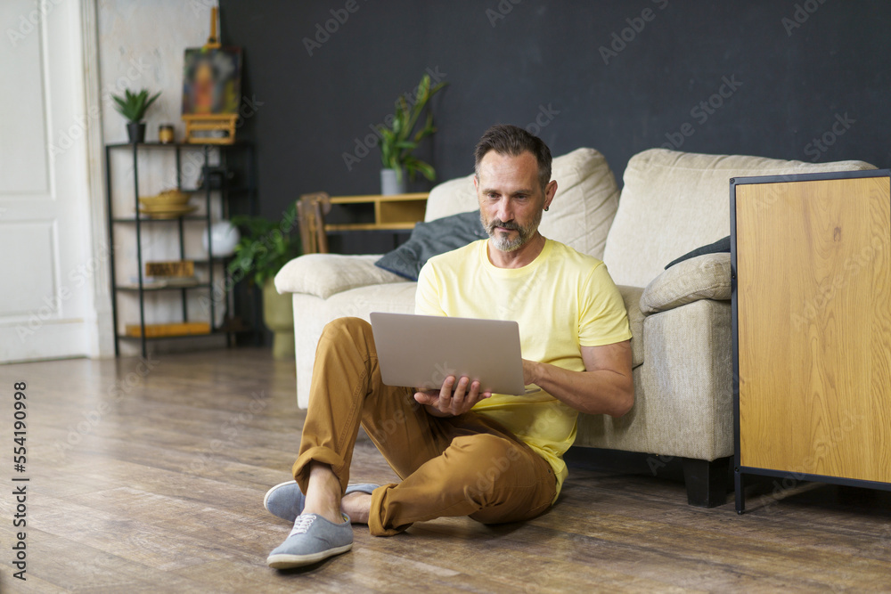 Fototapeta premium People and Technology Concept. Handsome mature man using laptop sitting on floor indoor lean on sofa watching video, movie or webinar, have a break