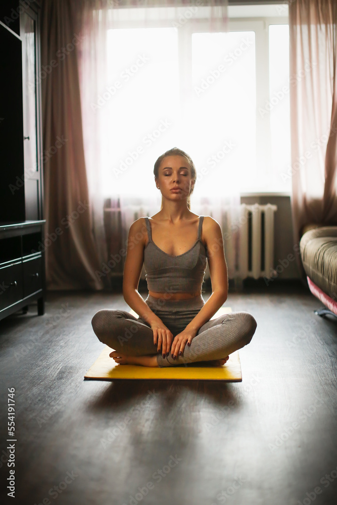 Woman with long hair extensions and large silicone lips. Pretty woman in gray sports top and leggings in lotus position meditates at home. Woman with long hair extensions and large silicone lips. Pretty woman in gray sports top and leggings in lotus position meditates at home.