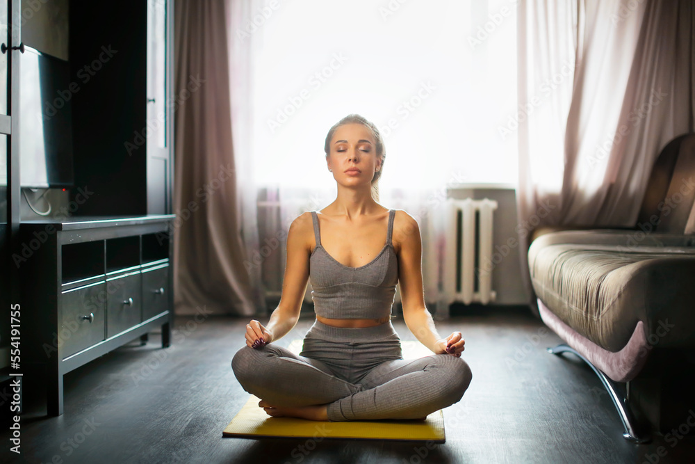 Pretty woman in gray sports top and leggings in lotus position meditates at home. Young woman does yoga on mat at home in living room . Pretty woman in gray sports top and leggings in lotus position meditates at home. Young woman does yoga on mat at home in living room .