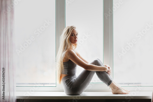 Slender young woman in gray tight tracksuit with long hair sits on window in room.