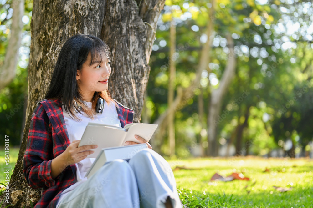Happy Asian female reading a book, daydreaming about her success while sitting under the tree Happy Asian female reading a book, daydreaming about her success while sitting under the tree