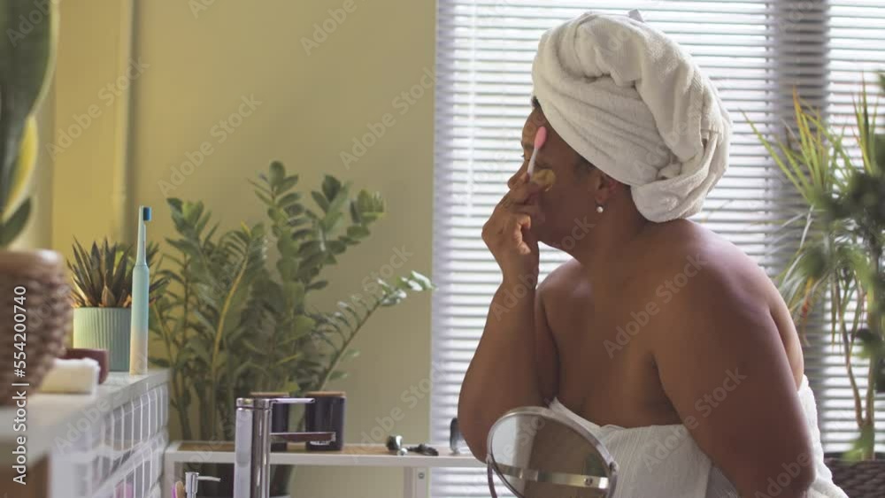 Smiling middle aged African American woman using silicone facial cleansing massager while doing skincare routine in bathroom after shower Smiling middle aged African American woman using silicone facial cleansing massager while doing skincare routine in bathroom after shower