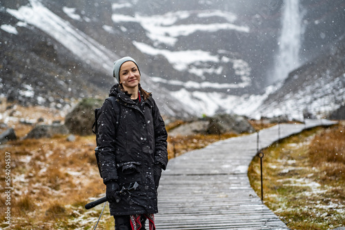 youn woman smiling, looking in camera and holding a camera in hand on stick on wooden trail at Hengifoss waterfall with layers of red clay during falling snow