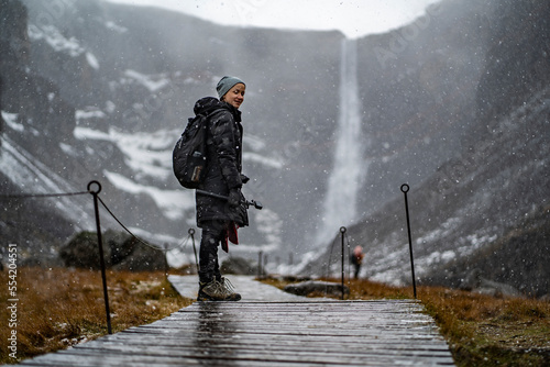 youn woman smiling, looking in camera and holding a camera in hand on stick on wooden trail at Hengifoss waterfall with layers of red clay during falling snow