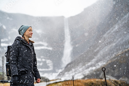 youn woman smiling, looking in camera and holding a camera in hand on stick on wooden trail at Hengifoss waterfall with layers of red clay during falling snow