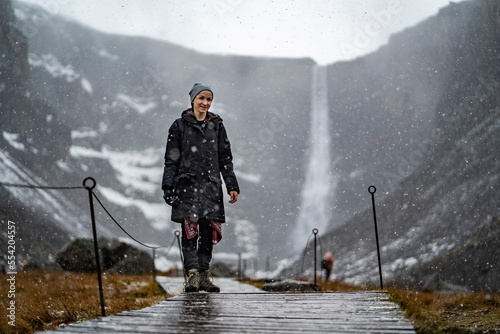 youn woman smiling, looking in camera and holding a camera in hand on stick on wooden trail at Hengifoss waterfall with layers of red clay during falling snow