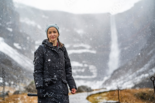 youn woman smiling, looking in camera and holding a camera in hand on stick on wooden trail at Hengifoss waterfall with layers of red clay during falling snow