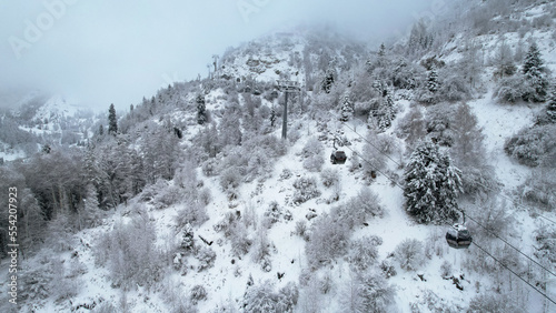 Cable car in the mountains in the winter forest. The cabins move along the gondola road through the winter snow forest and fog. White clouds covered the mountains. Top view from a drone. Medeo, Almaty