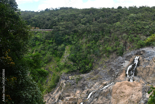 The white foam of a waterfall is stark against a cliff face. A forest is in the background. The red and white carriages of a train are visible in the distance.