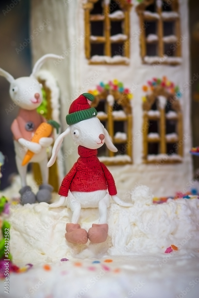 Christmas sugar figurine of a hare boy in a red sweater and hat on the background of a gingerbread house Christmas sugar figurine of a hare boy in a red sweater and hat on the background of a gingerbread house
