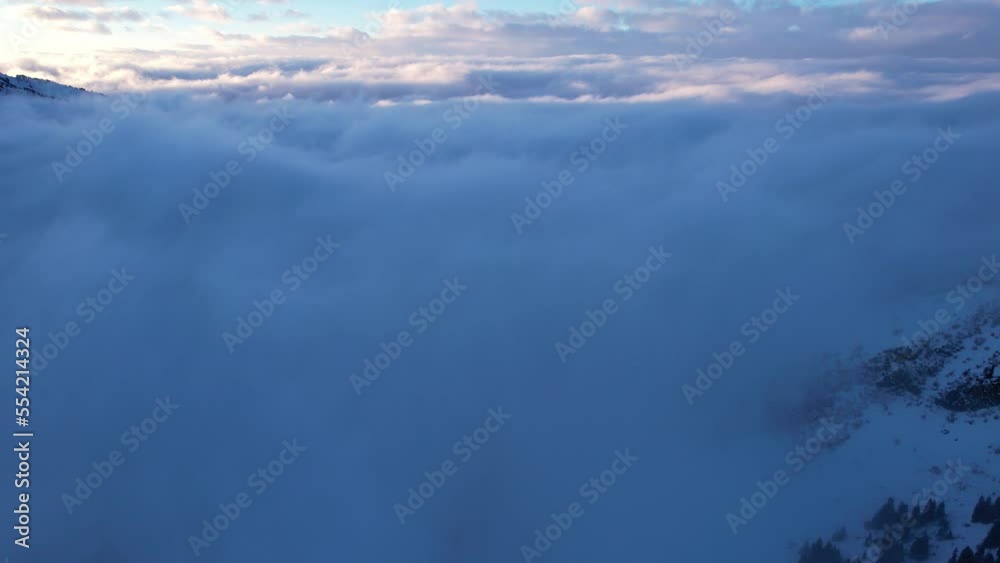 Flying among the ocean of clouds in the mountains. The fog is like big ocean waves crashing against high mountains covered with snow. Spruce trees grow in places. Sunset. Orange rays of the sun