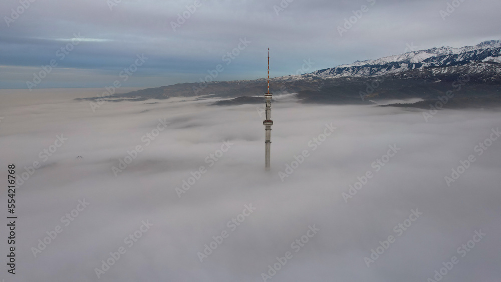The TV tower looks out of the clouds at sunset. Top view from a drone on a double layer of clouds. Yellow-blue shade of clouds. Shadows on clouds. High mountains are visible in the distance. Kok Tobe