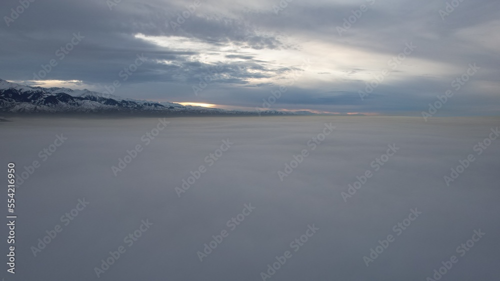 Flying a drone among the clouds with a view of the mountains. The light yellow rays of the sun at sunset are reflected on the surface of the clouds. Double layer of clouds. Mountains are visible
