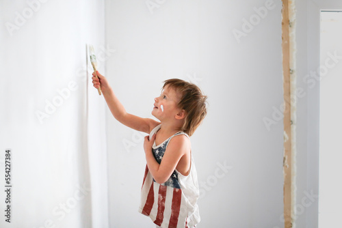 Cute little toddler girl painting wall with white color in new house.. Copy space. amily repair apartment home. Happy child girl paints the wall photo