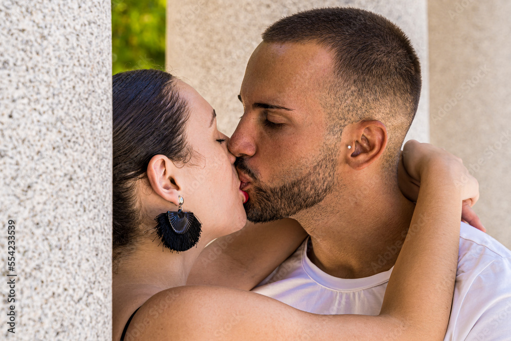 Fototapeta premium Close-up shot of a young couple kissing between stone columns, wearing black earrings and white shirt.