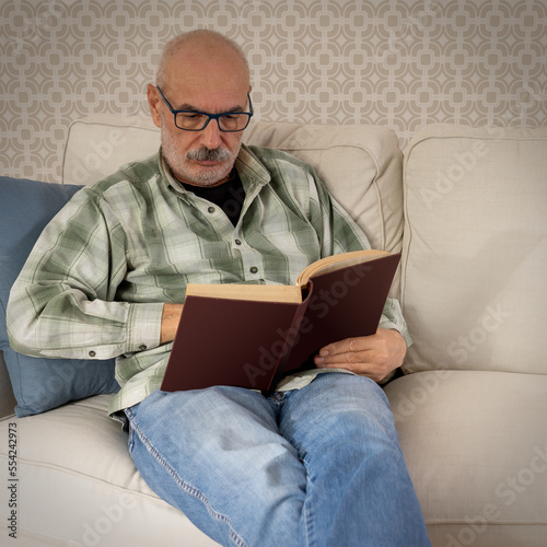 Happy Senior Man Sitting on Sofa and Reading Book at Home