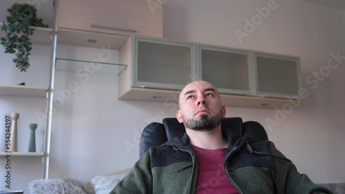 Young Caucasian bearded bald man sitting on chear and thinking. Depressed person in living room. Psychology and mental health. photo