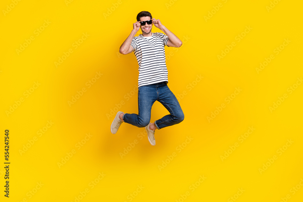 Full length photo of funny cool guy dressed striped t-shirt dark eyewear jumping high having fun isolated yellow color background
