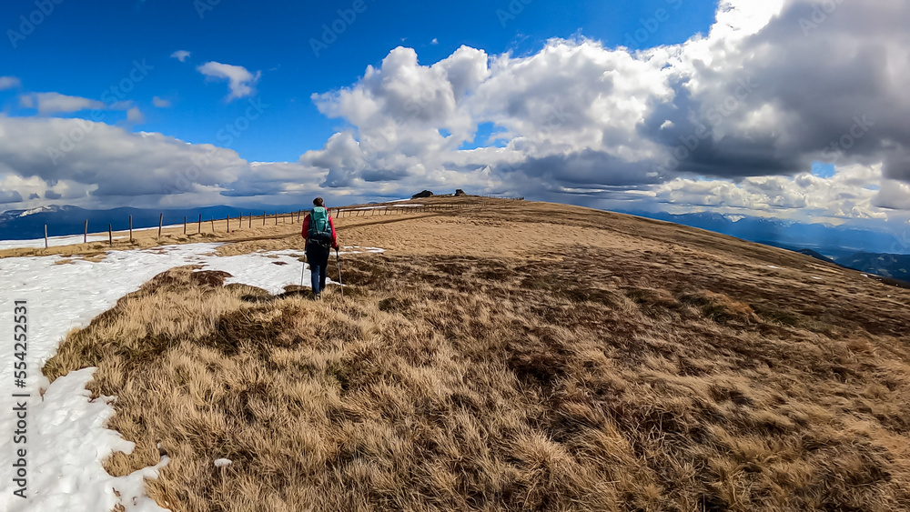 Woman with backpack on hiking trail walking next to fence on alpine meadow, Saualpe, Lavanttal Alps, Carinthia, Austria, Europe. Trekking on early spring day. View on valley of Wolfsberg. Freedom