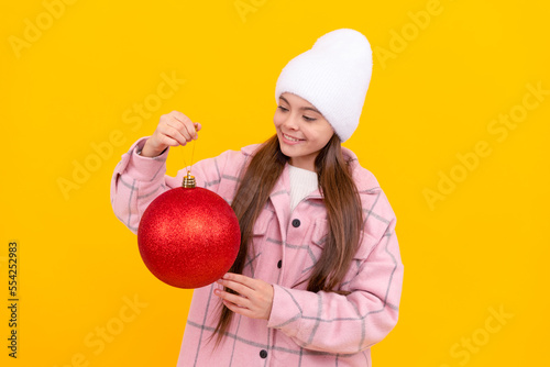 cheerful kid in winter hat hold christmas decorative ball on yellow background, new year