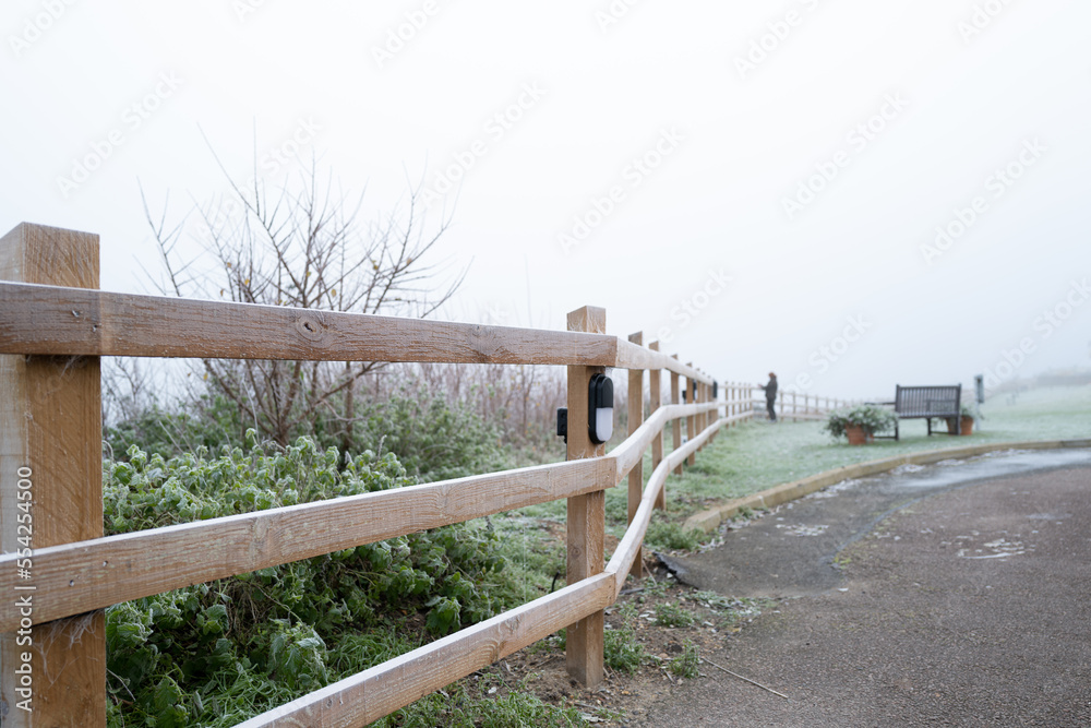 Shallow focus of a wooden fence sent on a wintery morning with frost settled. A person in the distance is looking out to the ocean which is covered with fog. Shallow focus of a wooden fence sent on a wintery morning with frost settled. A person in the distance is looking out to the ocean which is covered with fog.