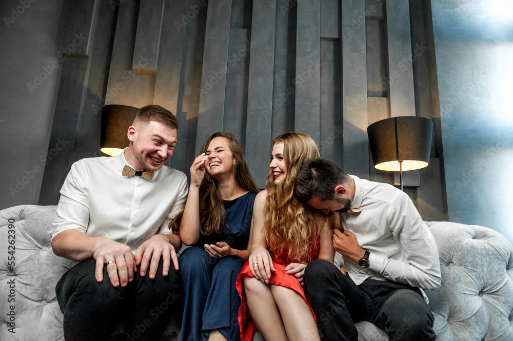 Obraz premium Friends in festive attire are sitting with glasses of wine on a sofa in a studio; on a gray sofa with a model posing; sitting close to each other smiling and grimacing at the camera