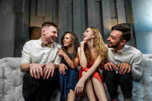 Friends in festive attire are sitting with glasses of wine on a sofa in a studio; on a gray sofa with a model posing; sitting close to each other smiling and grimacing at the camera