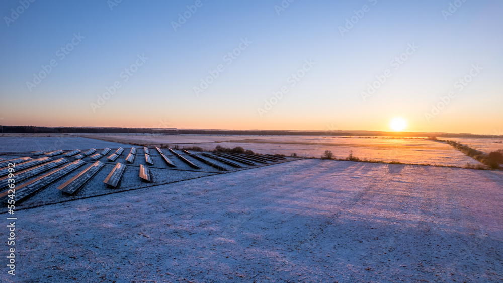 Aerial view of the solar farm during winter sunset. Blue sky, orange sun. Flight low over the white fields. 