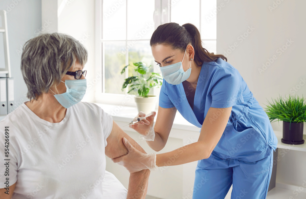 Doctor giving an arm injection to a senior woman. Young nurse in blue scrubs and surgical face mask holding a syringe and giving a vaccine shot to a retired old lady. Vaccination, immunization concept - obrazy, fototapety, plakaty 