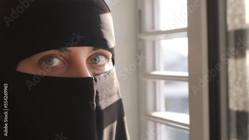 Muslim woman all covered in black looking out the window for freedom. Sad woman with Hijab and Niqaab at home. Light shining on her face. Hamas