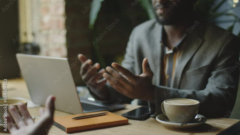 Cropped shot of African American businessman sitting with laptop and cup of coffee at cafe table, discussing construct with partner and giving him handshake Cropped shot of African American businessman sitting with laptop and cup of coffee at cafe table, discussing construct with partner and giving him handshake