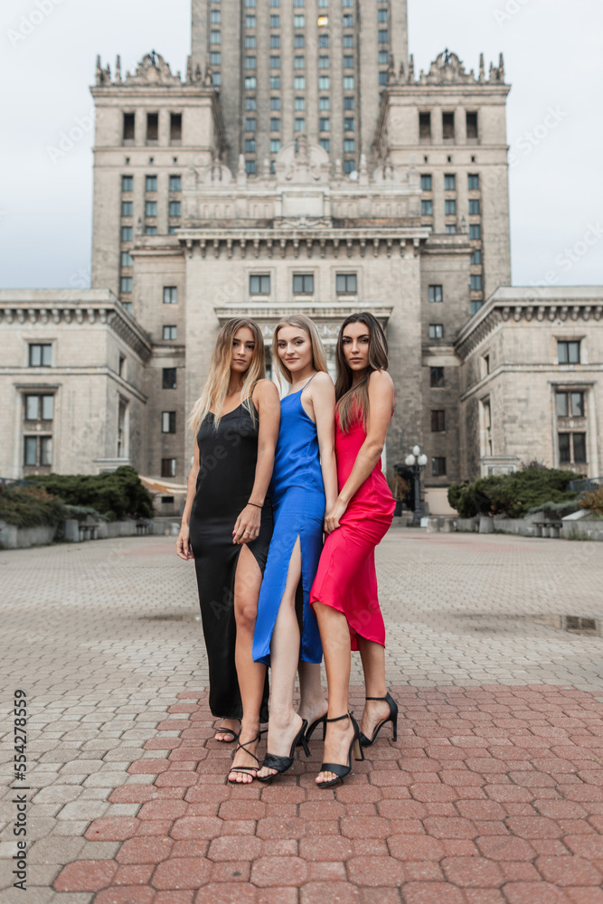 Three beautiful elegant slender girl friends in a fashionable dress with heels shoes stand in the city