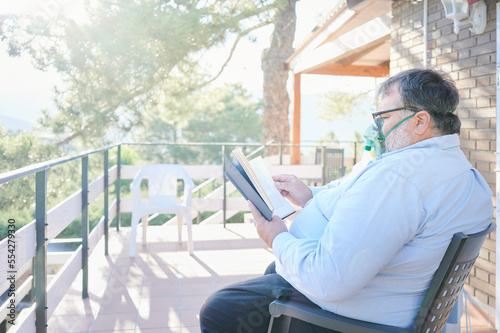 Overweight senior man patient recovers from his respiratory disease using a portable oxygen concentrator while reading a book on the balcony of his house. Aftermath after suffering from COVID-19. photo