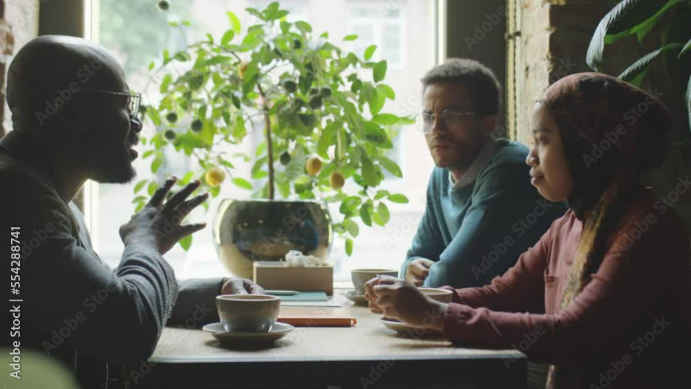 Multiethnic businessmen and Muslim businesswoman in hijab drinking coffee and talking while meeting in cafe Multiethnic businessmen and Muslim businesswoman in hijab drinking coffee and talking while meeting in cafe