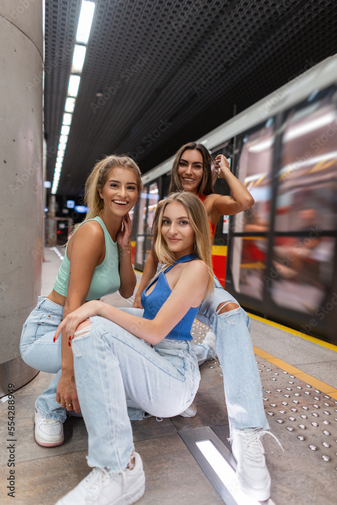 Happy beautiful young stylish women friends with smile in fashionable street clothes with a top, blue jeans and white sneakers are sitting and having fun on the subway near the moving train Happy beautiful young stylish women friends with smile in fashionable street clothes with a top, blue jeans and white sneakers are sitting and having fun on the subway near the moving train