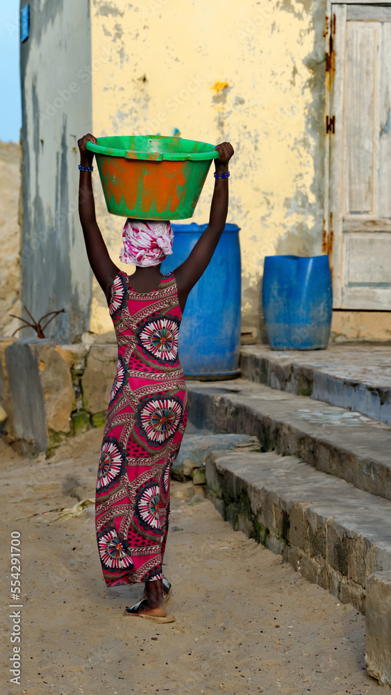 West Africa. Senegal. A young girl in national clothes gracefully carries a plastic basin with a load on her head along the street of the city of Saint-Louis.